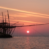 August 2016  Trabucci sind Pfahlbauten der italienischen Fischer an der apruzzesischen Adriaküste und entlang der Halbinsel Gargano. Ich verbinde die Trabucci mit unserem Pfingst- und Sommerdomizil dem Spiaggia Scialmarino bei Vieste. Am nördlichen Ende der Bucht auf der Insel La Chianca befindet sich ein großes, noch genutztes Trabucco. Ich habe mir vorgenommen, es mit der aufgehenden Sonne im Hintergrund zu fotografieren. Um Rechtzeitig vor Ort zu sein, musste ich ganz entgegen meiner sonstigen Gepflogenheiten, sehr früh aufstehen. Auf dem Hinweg habe ich mich noch über die vielen Kondensstreifen am Horizont geärgert, aber im nachhinein betrachtet wirken die diagonal verlaufenden und sich bereits auflösenden Kondensstreifen sogar gut …